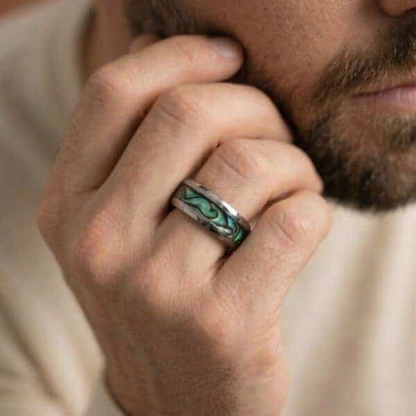 A man with a beard sits in a thoughtful way. He has his hand to his beard so you can see his magnificent tungsten ring. This silver ring has an inly of abalone shell. The edges of the ring are multi faceted so catch the light from all around.