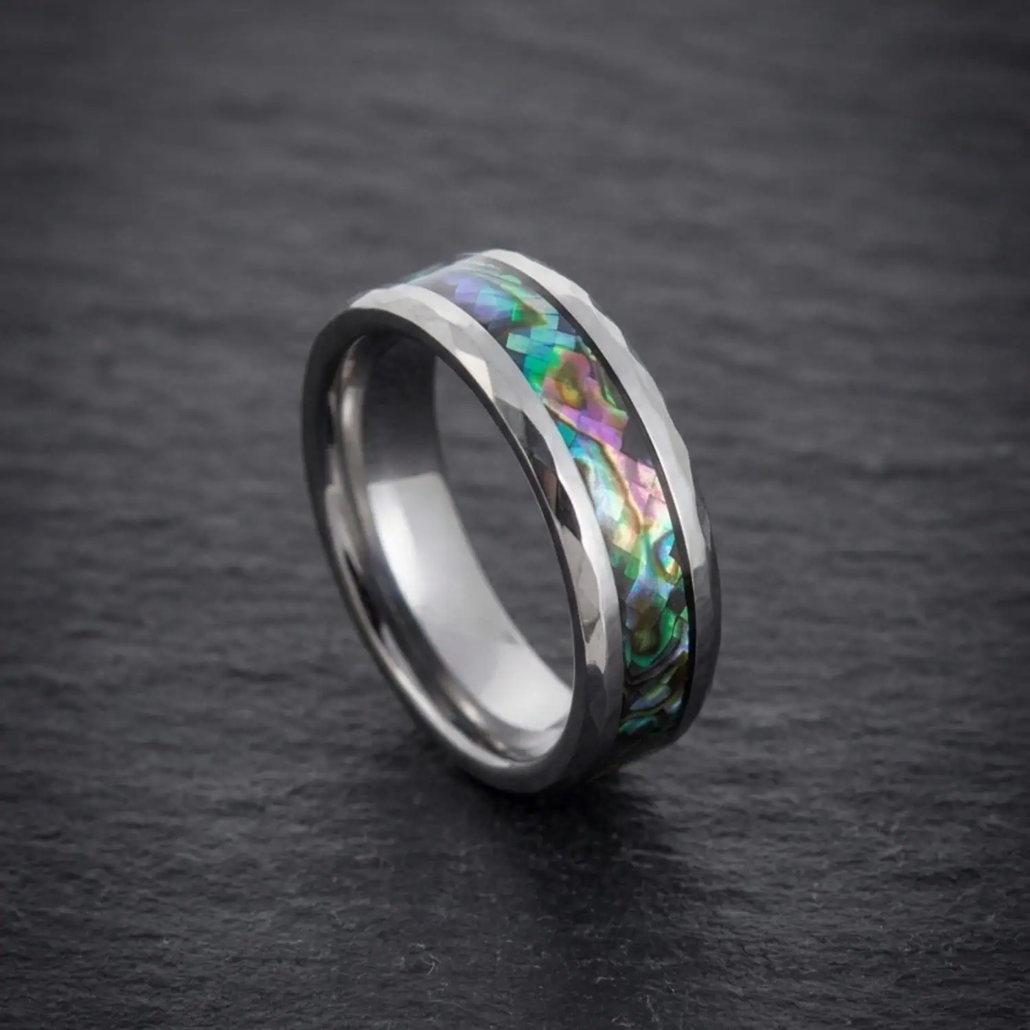 Reef silver tungsten carbide ring standing upright on a slate surface, highlighting the shimmering abalone shell inlay and multi-faceted bevelled edges.
