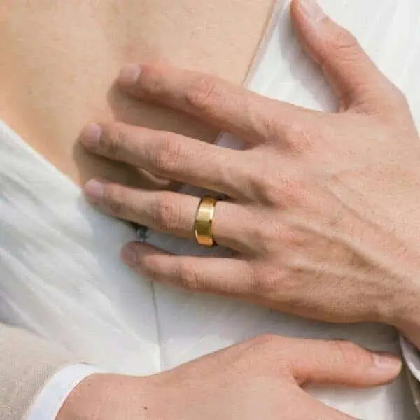 A close up image of a bridge and groom as they hold each other. You can see the back of the bride while the man has his arms around her. You can see his hands in detail. He is wearing a gold tungsten ring. It has a brushed surface and polished bevelled edges.