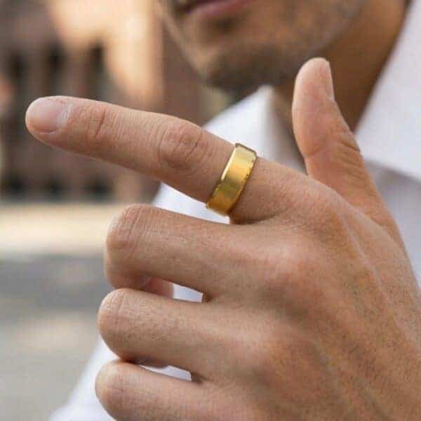 A zoomed in image of a mans hand with a city scene in the background. The man has his hand raised in front of him. He is wearing a gold tungsten ring on his index finger. It is a gold ring that features brushed and polished sections. It has a flat section which has bevelled edges.