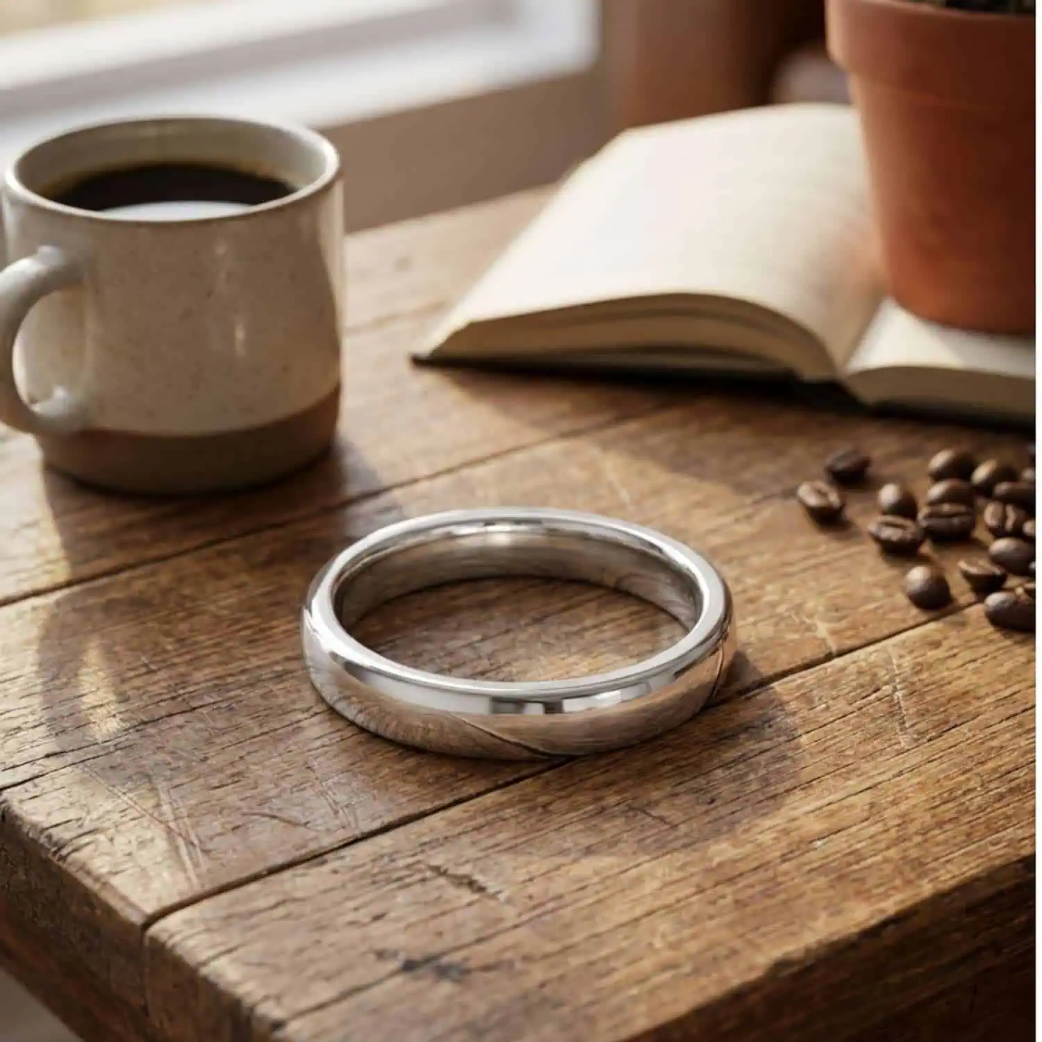 Legacy polished white gold tungsten ring resting on a coffee table beside a cup of coffee, coffee beans and an open book.