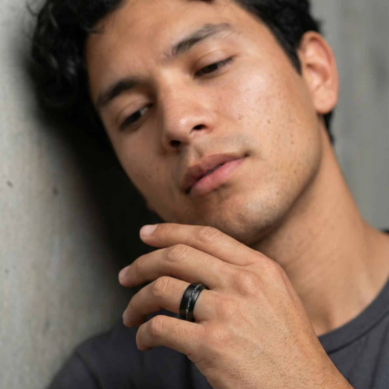 The young man leans against a wall while raising his hand. He is wearing a brushed black tungsten ring that has an inlay of faux crushed meteorite.
