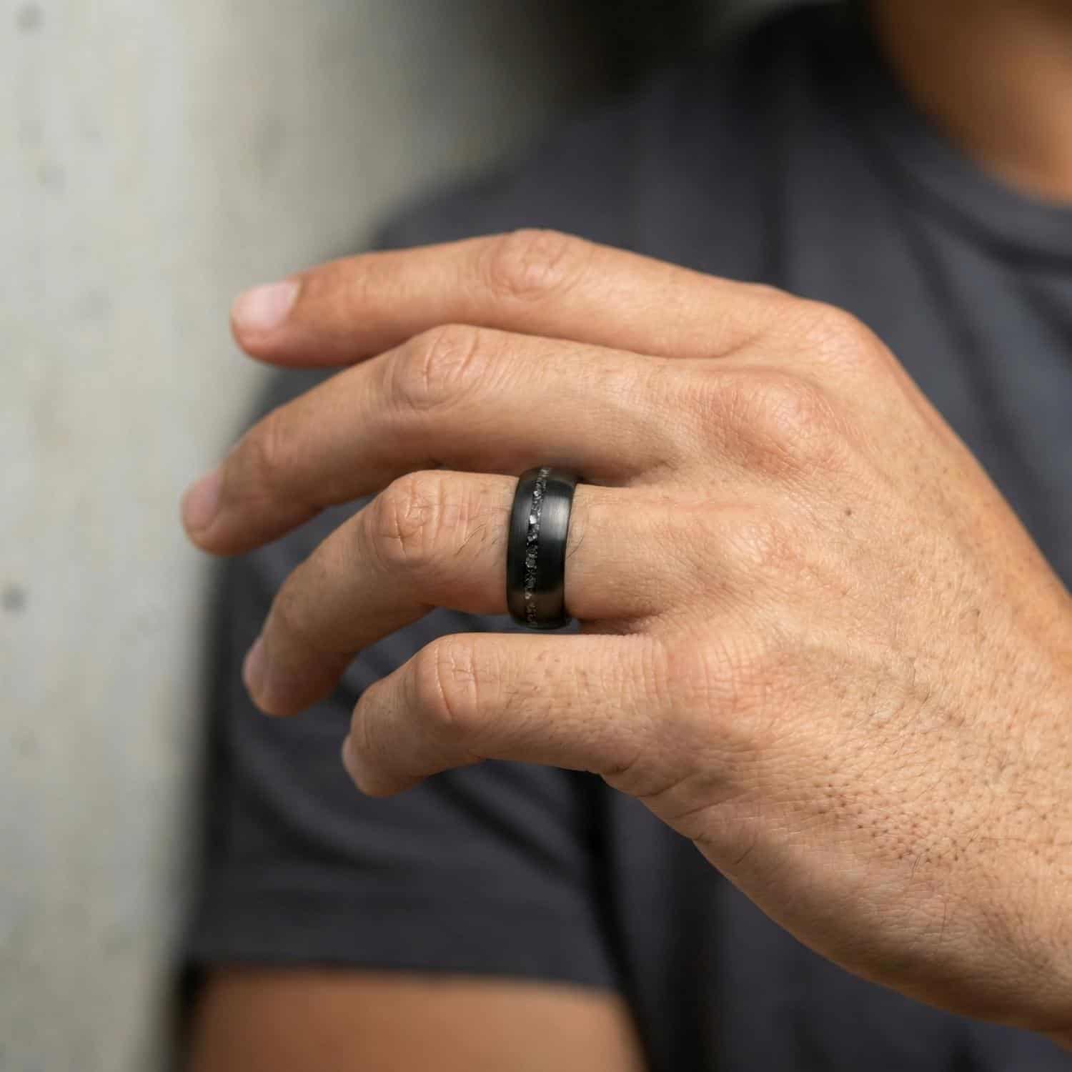 A close up of a mans hand. He is wearing a brushed black tungsten ring that has a groove of faux crushed meteorite.