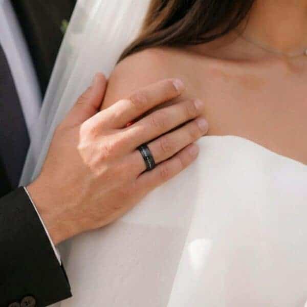 At the wedding ceremony, the man holds the shoulder of the bride. He is wearing a brushed black tungsten ring that features a groove of faux crushed meteorite.