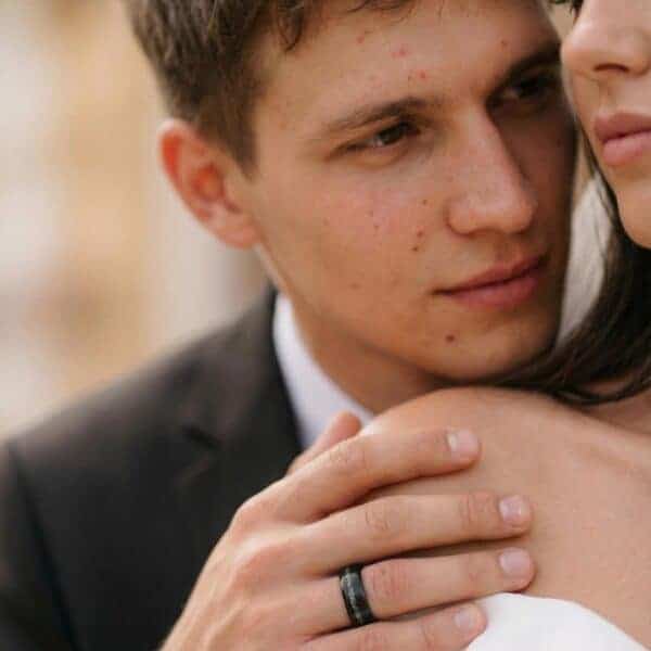 The wedding couple share an embrace. The mans hand is visible, showing his black tungsten ring that has a brushed finish and an inlay of faux crushed meteorite.