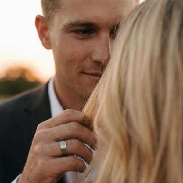 A newly married couple face each other, the man facing the camera. Reaching out to touch her, he raises his hand. On his finger is a white gold tungsten ring.