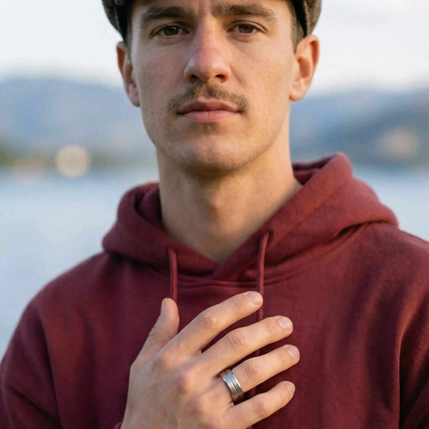 Man wearing a hoodie standing outdoors by a lake while wearing the Zenith men's white gold tungsten ring with a polished centre groove and brushed raised sections.