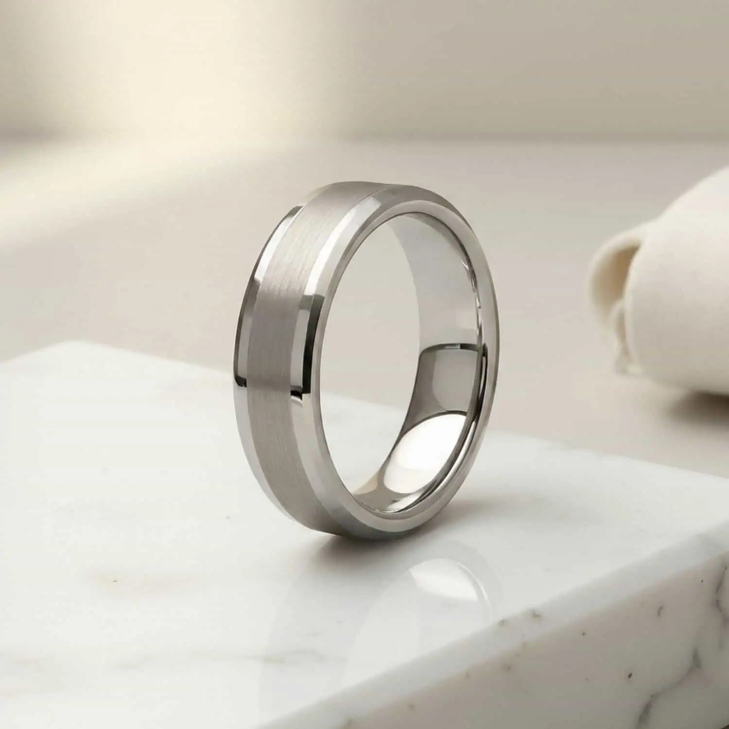 A white gold tungsten ring standing upright on white marble, showcasing the brushed centre, polished sections, and polished bevelled edges against a minimalist white background.