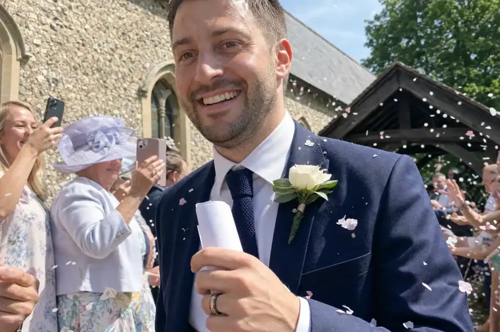 A smiling groom in a navy suit with a white rose buttonhole walking out of a flint village church through falling confetti, holding a rolled order of service, the Champion tungsten ring on his ring finger