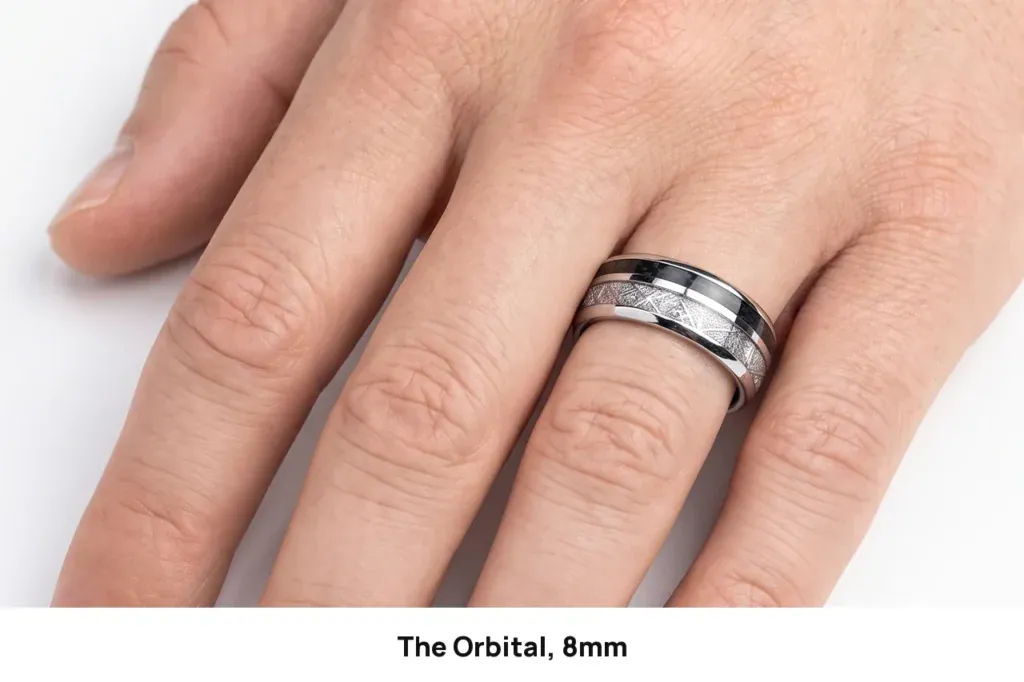 The Orbital silver tungsten ring with its meteorite-pattern inlay and carbon-fibre stripe, worn on the ring finger of a hand against a white studio background