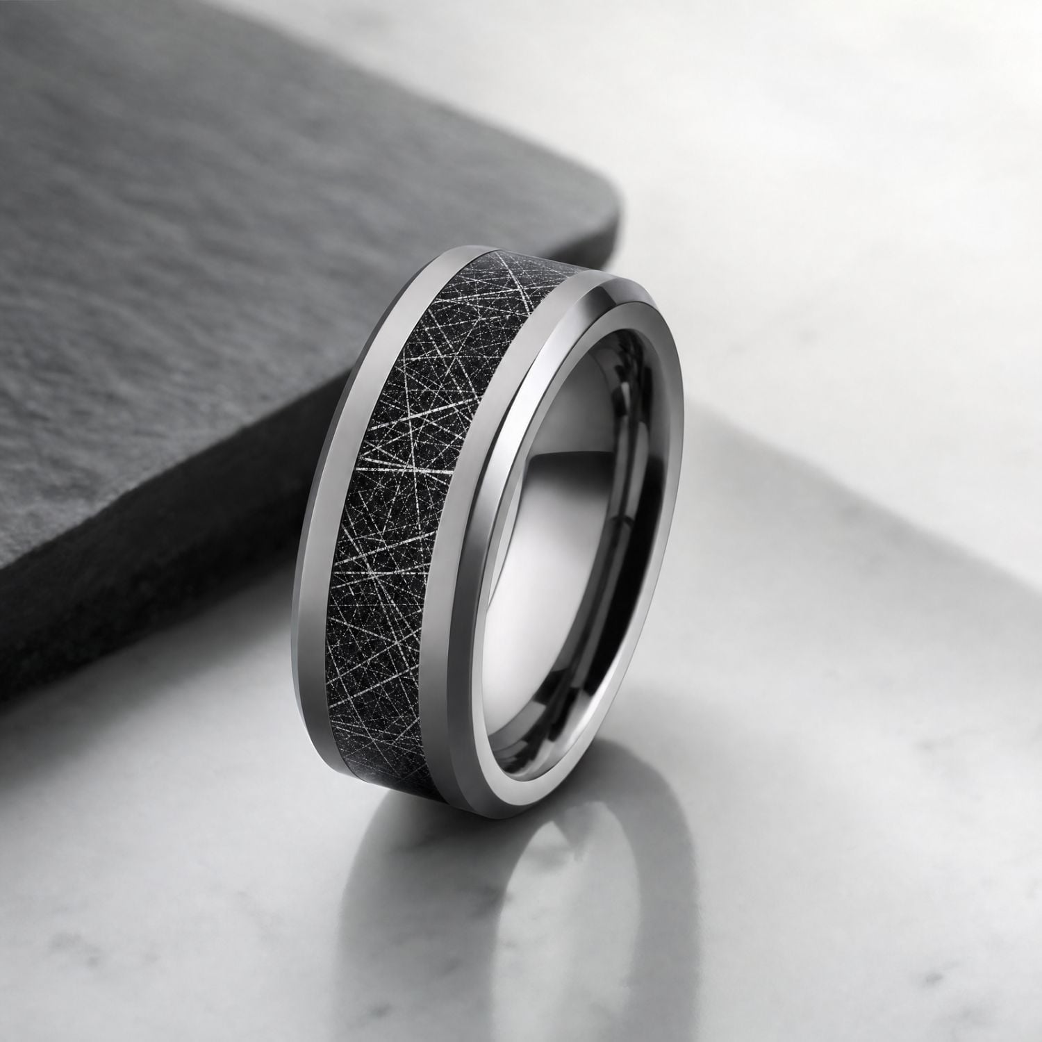 Silver tungsten ring standing upright on a smooth light grey surface beside charcoal slate, highlighting its black faux meteorite inlay and polished bevelled edges.