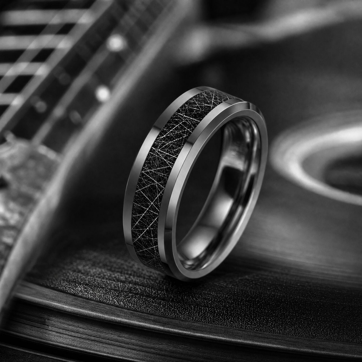 Silver tungsten ring with a black faux meteorite inlay standing upright on a vinyl record, with guitar strings visible along the edge of the music-themed image.