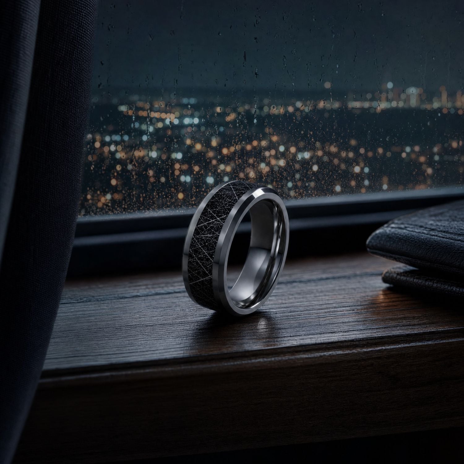 Men's polished silver tungsten ring with a black faux meteorite centre standing on a dark wooden windowsill, overlooking distant town lights beneath the night sky.