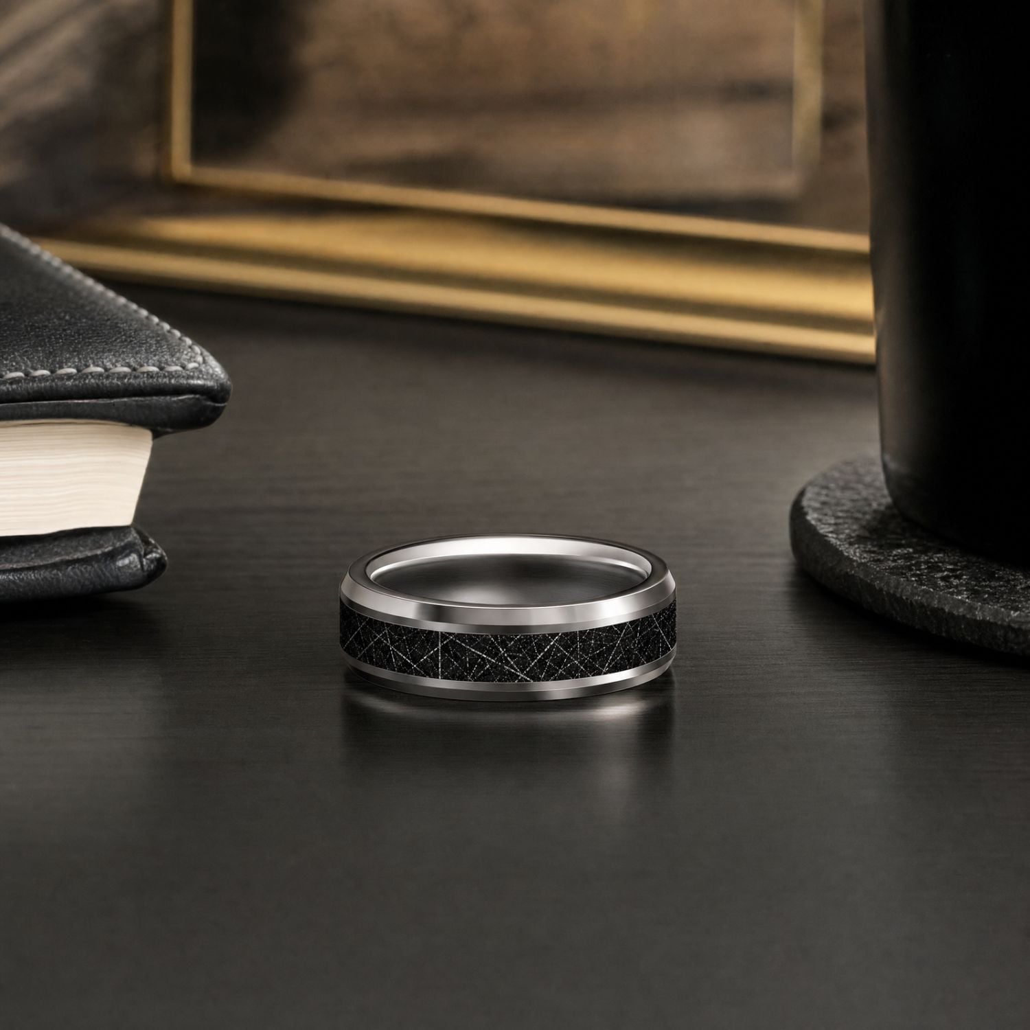 Men's silver tungsten ring with a black faux meteorite centre resting on a dark wooden shelf beside a leather-bound book, with a gold-framed mirror and lamp behind it.