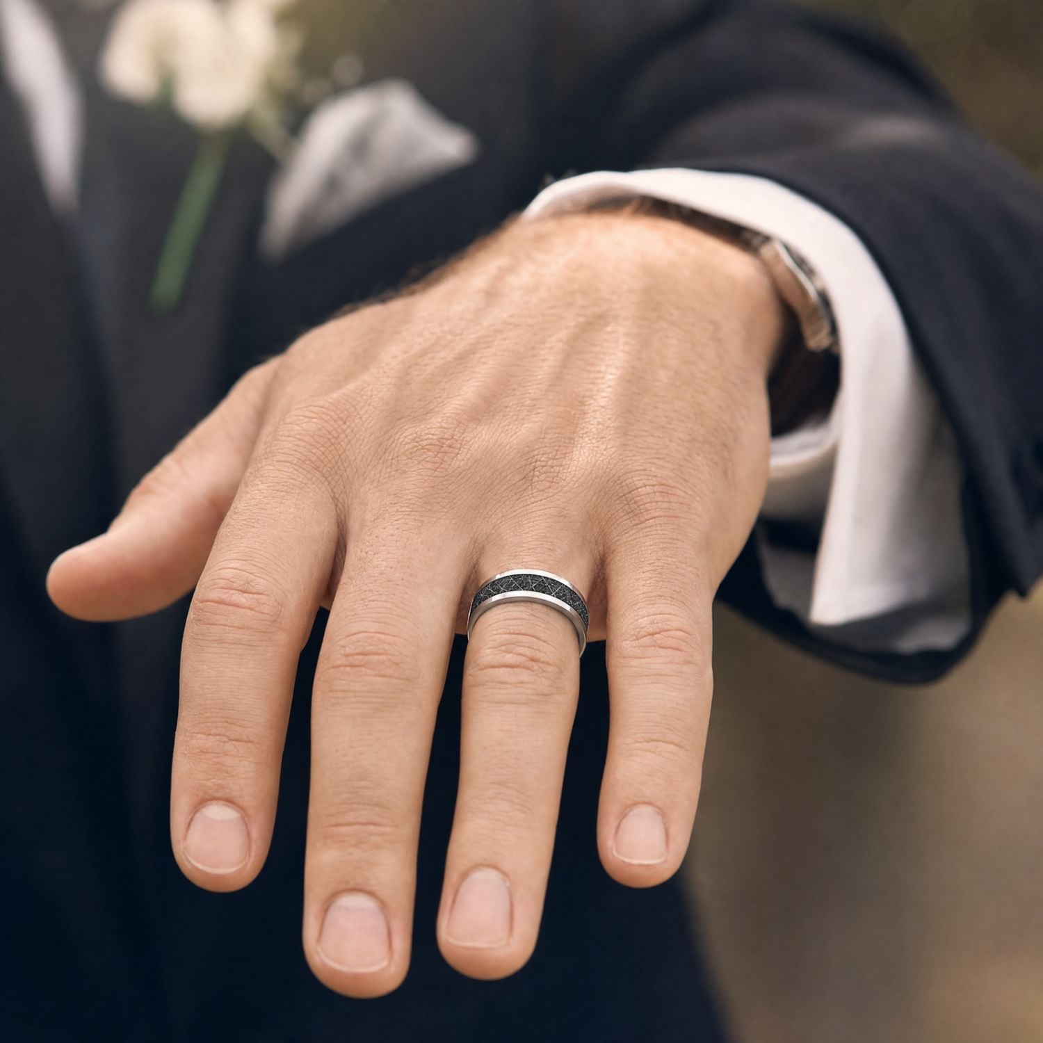 Groom in a dark suit with a wedding flower pinned to his jacket holding out his hand to show a silver tungsten wedding ring with a black faux meteorite inlay.