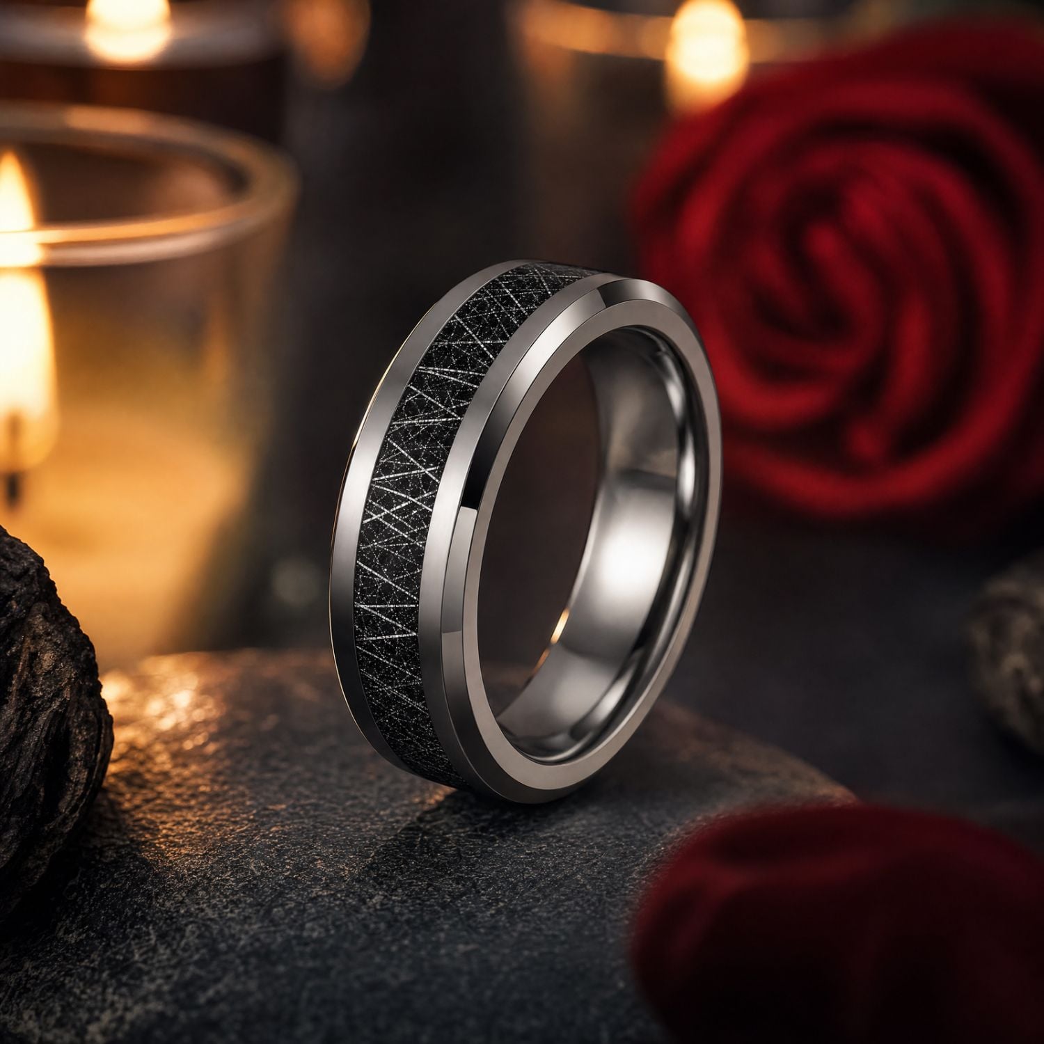 Polished silver tungsten wedding band with a black meteorite-style inlay standing on a rounded stone beside a red rose, with softly glowing candles in the background.