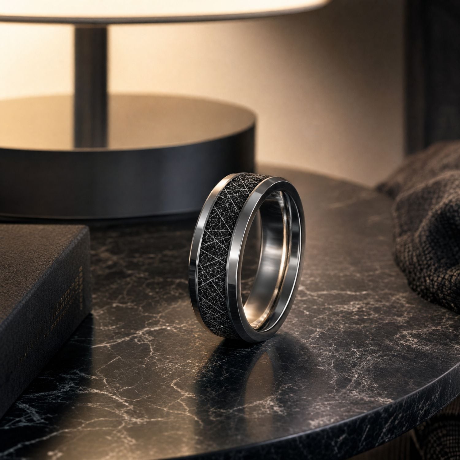 Polished silver tungsten ring with a black faux meteorite inlay standing on a marble bedside table beside a leather-bound book under the warm glow of a lamp.