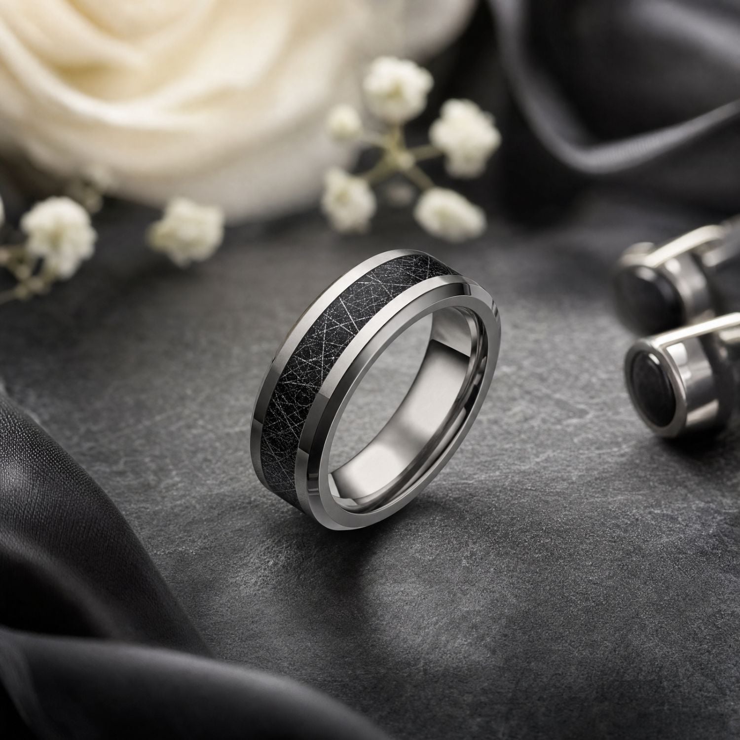 Polished silver tungsten wedding band with a black meteorite-style centre displayed beside silver and black cufflinks, white wedding flowers and a white rose.