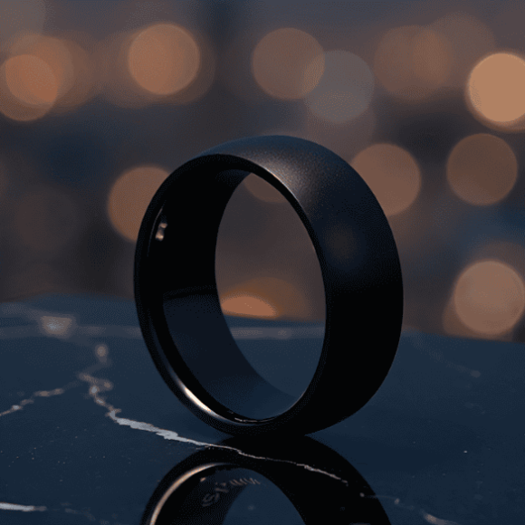Sleek black wedding band on a dark surface with bokeh lights in the background.